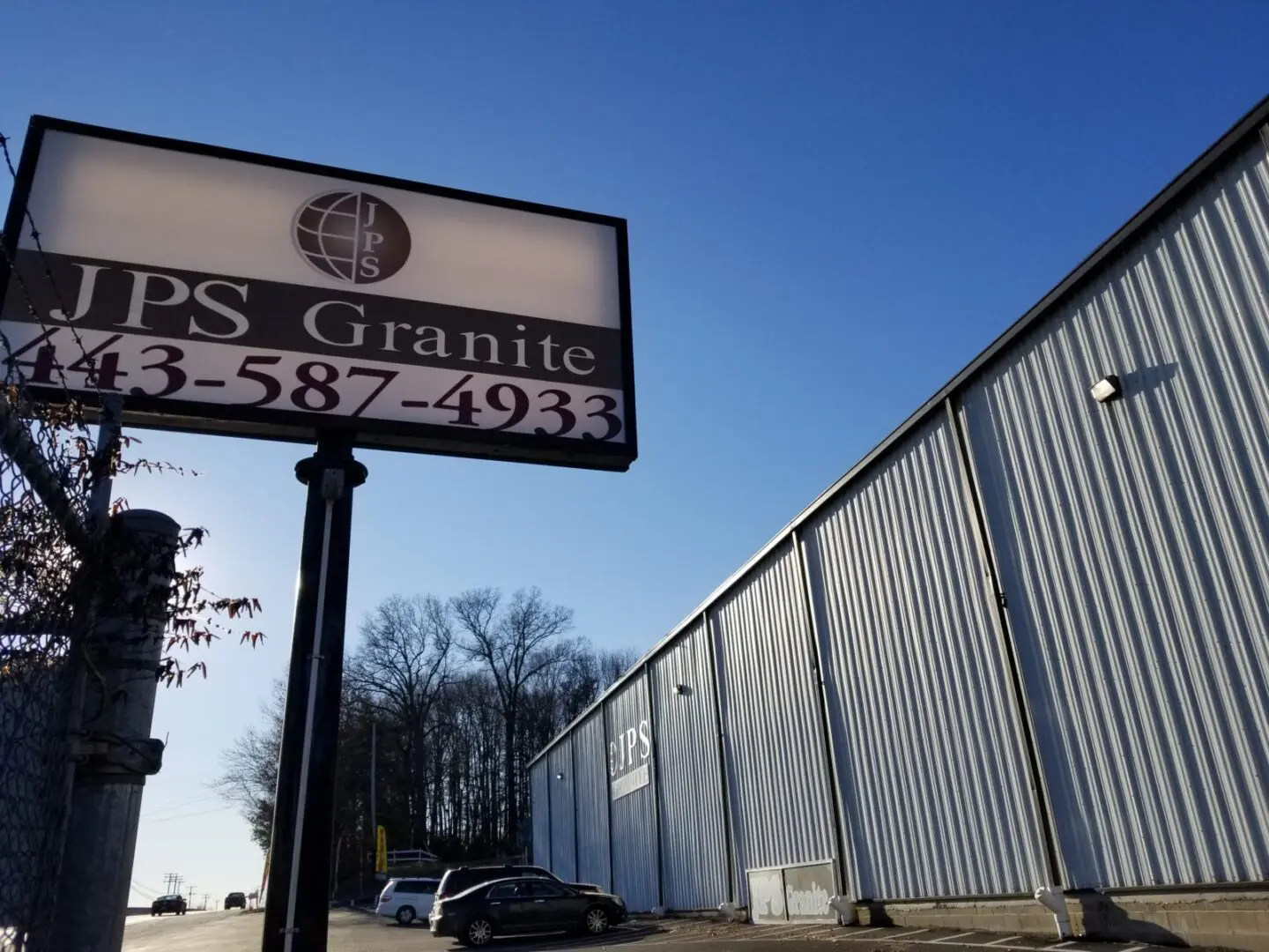 A sign for jps granite with contact information displayed in front of a corrugated metal building under a clear sky.