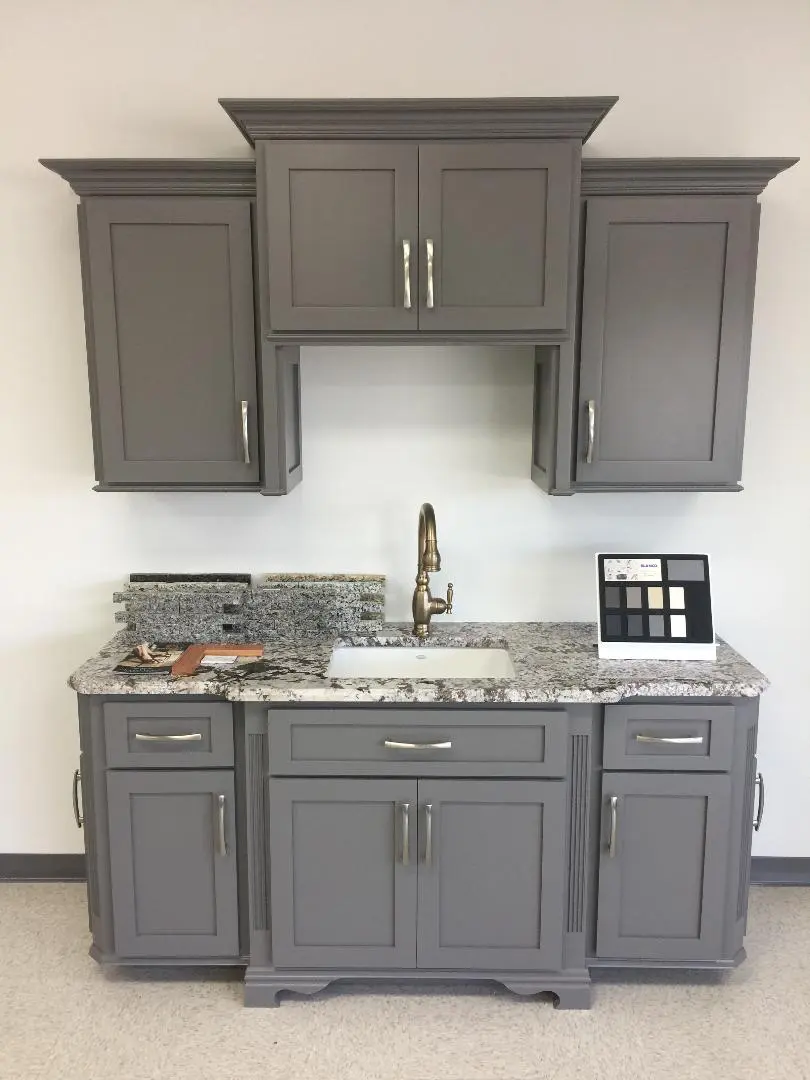 Modern grey kitchen cabinetry with granite countertop and a brass faucet, displayed in a showroom setting.