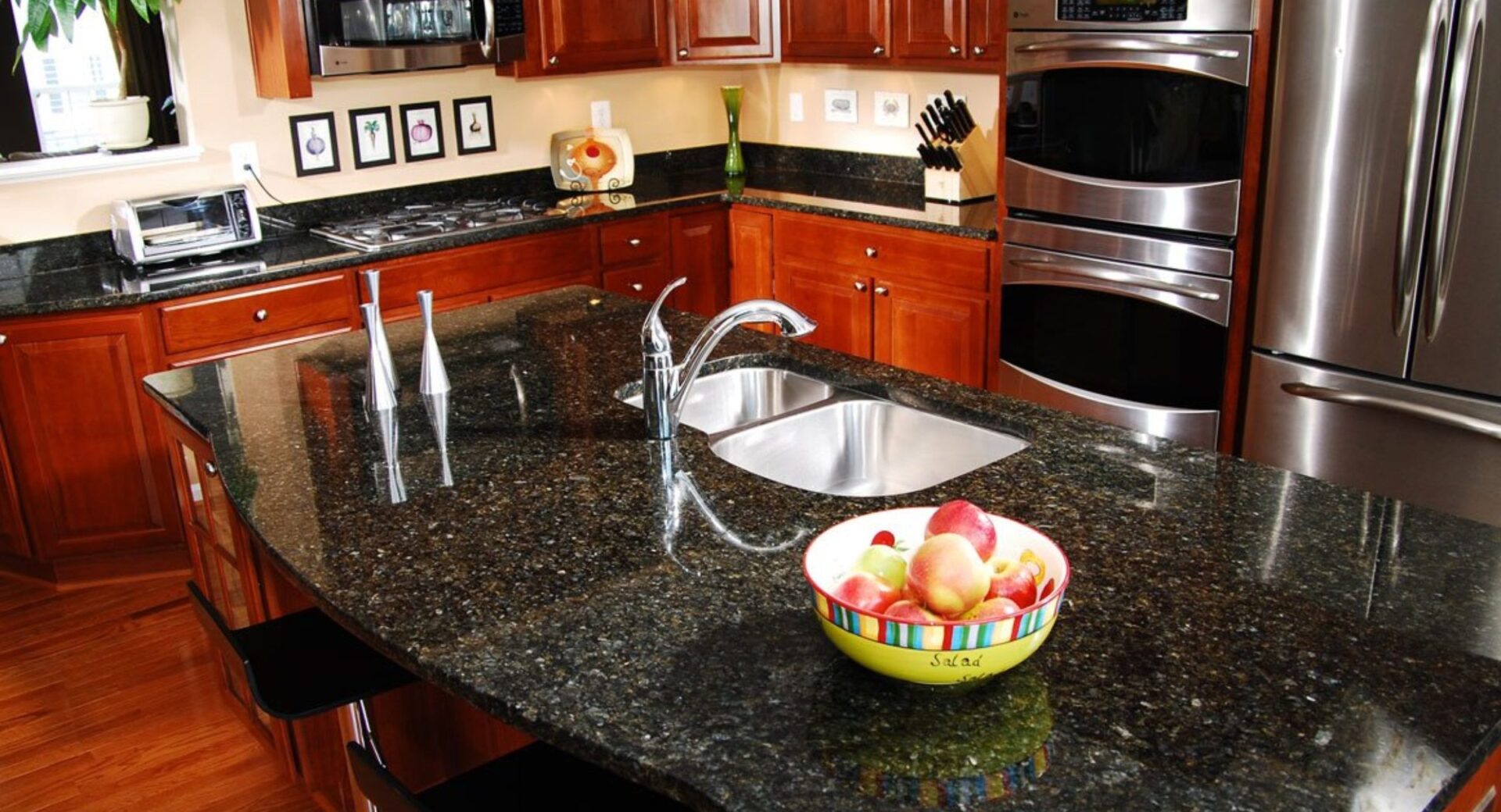 A modern kitchen with stainless steel appliances and a granite countertop, featuring a bowl of fruit on the island.